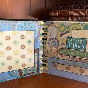 May include: Open scrapbook with patterned pages. The left page has a blue background with a large square of patterned paper featuring shamrocks and other designs. The right page has the text "EARNING BADGES" on a green background.