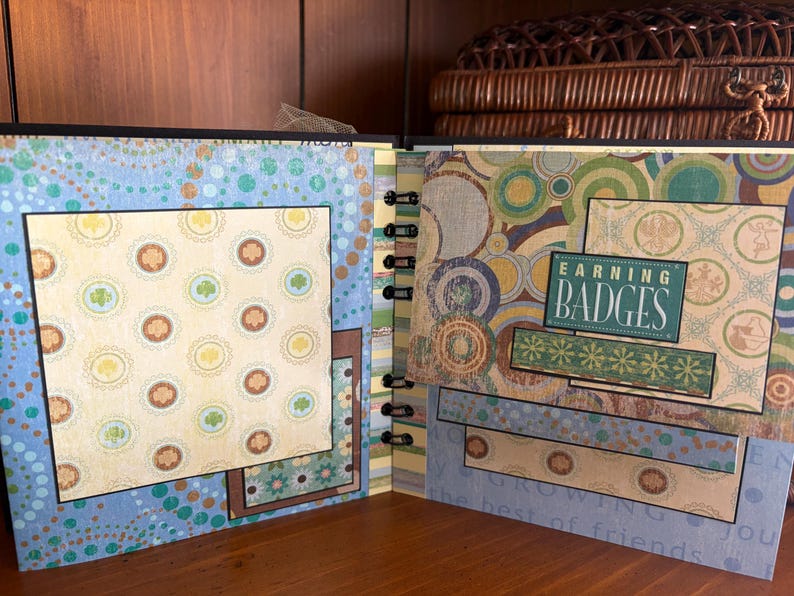 May include: Open scrapbook with patterned pages. The left page has a blue background with a large square of patterned paper featuring shamrocks and other designs. The right page has the text "EARNING BADGES" on a green background.
