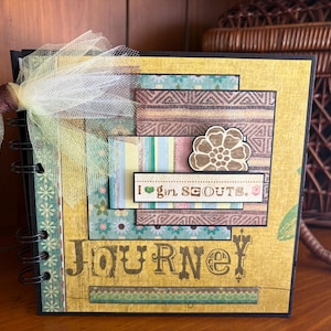 May include: A handmade scrapbook with a spiral binding. The cover is yellow with layered patterned paper in teal, brown, and blue. The text "JOURNEY" is printed on the front, along with the phrase "I girl SCOUTS."
