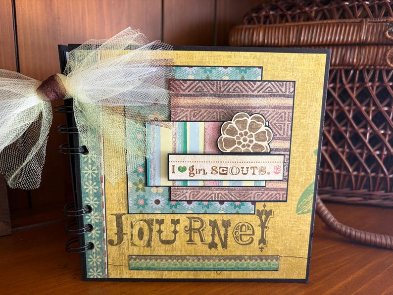 May include: A handmade scrapbook with a spiral binding. The cover is yellow with layered patterned paper in teal, brown, and blue. The text "JOURNEY" is printed on the front, along with the phrase "I girl SCOUTS."