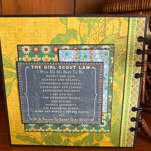 May include: A Girl Scout Law journal with a yellow floral background and a dark brown border. The journal features a layered design with a blue and green patterned insert. The text reads "The Girl Scout Law".