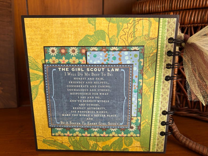 May include: A Girl Scout Law journal with a yellow floral background and a dark brown border. The journal features a layered design with a blue and green patterned insert. The text reads "The Girl Scout Law".
