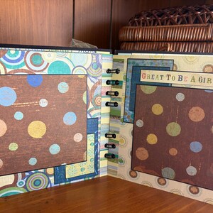 May include: Open scrapbook with brown, blue, and yellow polka dot pages. The scrapbook has a black spiral binding and a decorative cover with a colorful circular pattern. The text "GREAT TO BE A GIRL" is visible on one page.