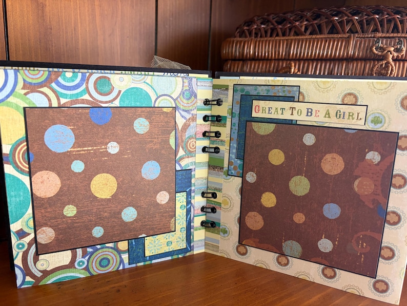 May include: Open scrapbook with brown, blue, and yellow polka dot pages. The scrapbook has a black spiral binding and a decorative cover with a colorful circular pattern. The text "GREAT TO BE A GIRL" is visible on one page.