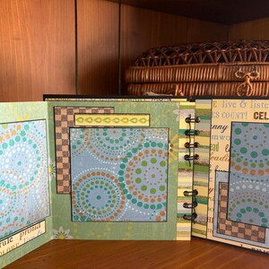 May include: Open scrapbook with pages featuring colorful dot patterns in teal, orange, and green. The pages are bound with a black spiral. Text includes "girls rule proud" and "CELEBRATE". The background includes a wooden shelf and a woven basket.