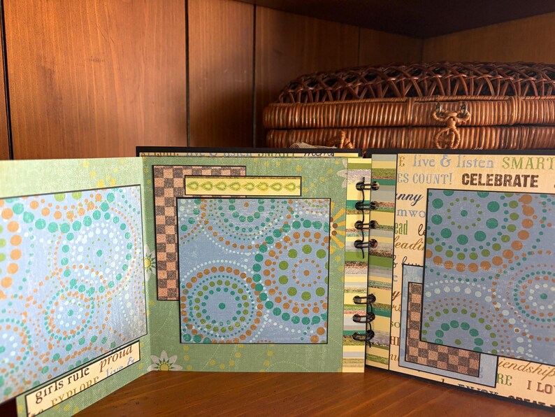 May include: Open scrapbook with pages featuring colorful dot patterns in teal, orange, and green. The pages are bound with a black spiral. Text includes "girls rule proud" and "CELEBRATE". The background includes a wooden shelf and a woven basket.