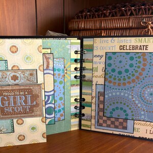 May include: An open scrapbook with pages featuring patterned paper and decorative elements. The cover displays a rectangular sign that reads "PROUD TO BE A GIRL SCOUT". The pages have a spiral binding and include text and colorful designs.