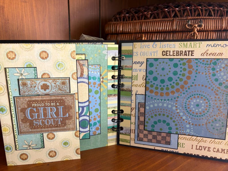 May include: An open scrapbook with pages featuring patterned paper and decorative elements. The cover displays a rectangular sign that reads "PROUD TO BE A GIRL SCOUT". The pages have a spiral binding and include text and colorful designs.