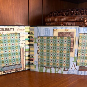 May include: Open scrapbook with floral patterned pages and decorative borders. The pages feature a mix of colors, including blue, green, and yellow, with text elements like "CELEBRATE" and "DREAM". The scrapbook is bound with black metal rings.
