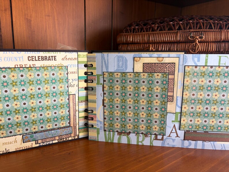 May include: Open scrapbook with floral patterned pages and decorative borders. The pages feature a mix of colors, including blue, green, and yellow, with text elements like "CELEBRATE" and "DREAM". The scrapbook is bound with black metal rings.