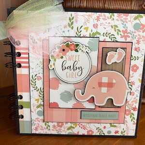 May include: A handmade baby girl scrapbook with a floral and gingham pattern. The cover features a pink elephant, baby footprints, and the words "Sweet Baby Girl" and "Welcome Home Baby."