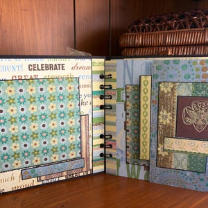 May include: Open scrapbook with floral and patterned paper pages. The left page has a floral square and text that reads "COOKIES COUNT! CELEBRATE". The right page features a brown square with a gold emblem.