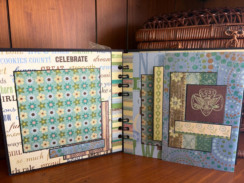 May include: Open scrapbook with floral and patterned paper pages. The left page has a floral square and text that reads "COOKIES COUNT! CELEBRATE". The right page features a brown square with a gold emblem.