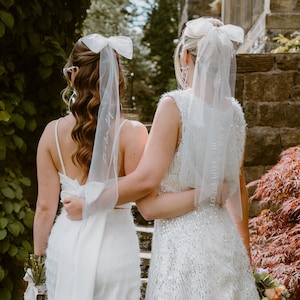 May include: Two people in white wedding outfits stand outdoors. One wears a white jumpsuit with a long veil and a bow. The other wears a short, sequined dress with a veil and bow. Both hold bouquets. The scene is set against greenery and a stone wall.