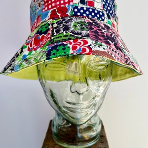 May include: A colorful patchwork bucket hat with a yellow brim. The hat is made of different fabrics with floral, geometric, and polka dot patterns. The hat is sitting on a glass head.