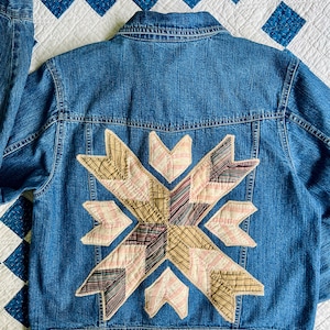 May include: A blue denim jacket with a large patchwork design on the back. The design is made up of various shades of brown, pink, and white fabric.
