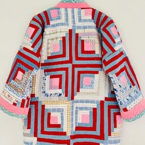 May include: A patchwork quilted jacket in a vibrant design. The jacket features a pattern of red, blue, and pink squares and rectangles. The sleeves and collar are trimmed with pink and green fabric. The jacket is hanging against a white backdrop.