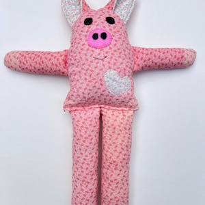 May include: A handmade pink pig stuffed animal with a heart-shaped patch on its chest. The pig has black button eyes, a pink snout, and floral patterned ears. The body and limbs are covered in a repeating pattern of pink shapes.