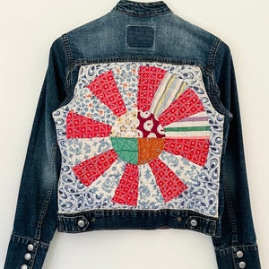 May include: A blue denim jacket with a patchwork design on the back. The patchwork is a circle of different colored fabrics with floral patterns.