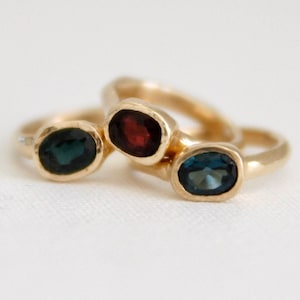 May include: Three gold rings with oval-shaped gemstones. The rings are stacked on top of each other. The top ring has a dark blue gemstone, the middle ring has a red gemstone, and the bottom ring has a light blue gemstone.
