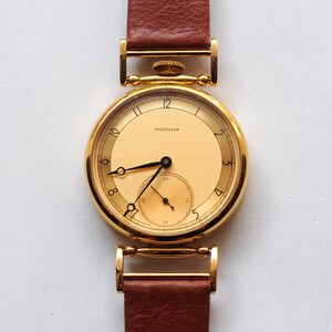 Può includere: Un orologio da polso dorato con un quadrante rotondo di colore crema e lancette nere. L'orologio ha un cinturino in pelle marrone e la parola "MORMAII" stampata sul quadrante.