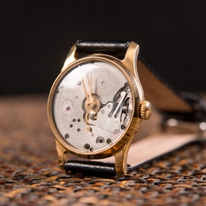 Peut inclure: Une montre-bracelet dorée avec un bracelet en cuir noir. Le cadran de la montre est ouvert, révélant les mécanismes internes, y compris des engrenages et un petit rubis. Les aiguilles indiquent environ 1h50. La montre est posée sur un fond sombre.