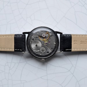 May include: A black leather wristwatch with a silver face and exposed gears. The watch band is beige with black stitching and the text "SK&Oacute;RA NATURALNA" printed on the band.