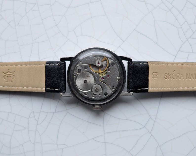 May include: A black leather wristwatch with a silver face and exposed gears. The watch band is beige with black stitching and the text "SK&Oacute;RA NATURALNA" printed on the band.