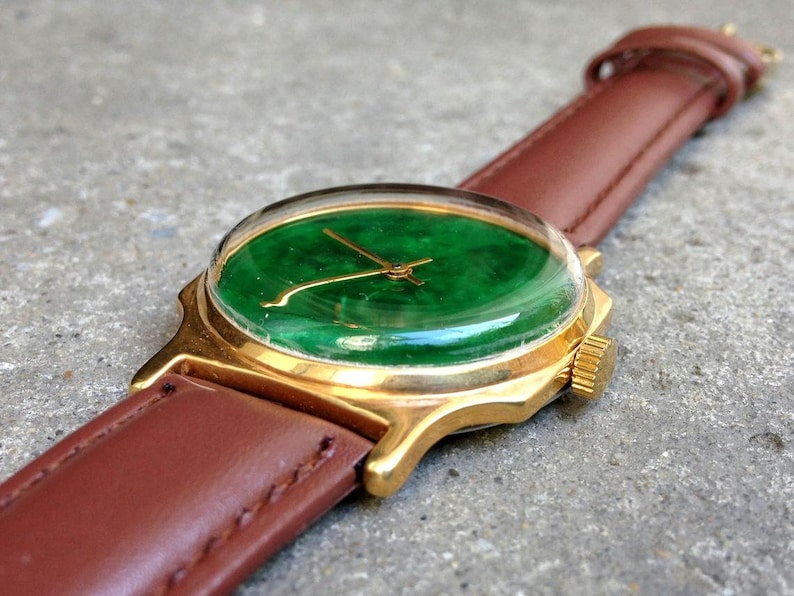 May include: A gold wristwatch with a green jade face and a brown leather band.