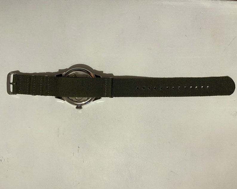 May include: A silver watch with a green nylon strap. The watch face is visible, but the time is not displayed.