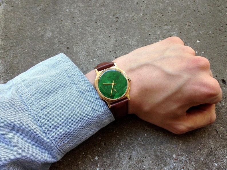May include: A gold-tone wristwatch with a green face and brown leather band.