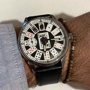 May include: A silver wristwatch with a black leather band. The watch face features a black background with playing cards arranged in a circle around the clock hands. The cards include the King of Spades, the Queen of Hearts, and the Jack of Diamonds.