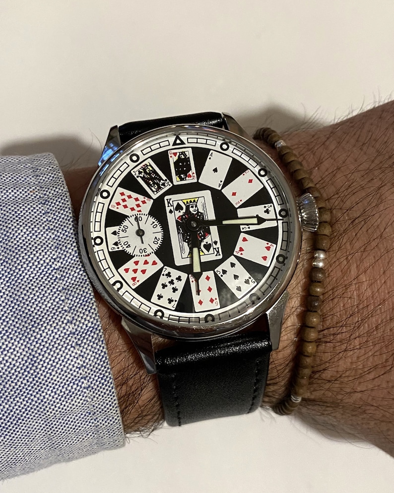 May include: A silver wristwatch with a black leather band. The watch face features a black background with playing cards arranged in a circle around the clock hands. The cards include the King of Spades, the Queen of Hearts, and the Jack of Diamonds.
