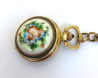 May include: A vintage gold-coloured pocket watch with a floral design. The watch face features a hand-painted floral motif in shades of orange, blue, and green on a white background. The watch has a decorative chain and a small, red jewel on the clasp.