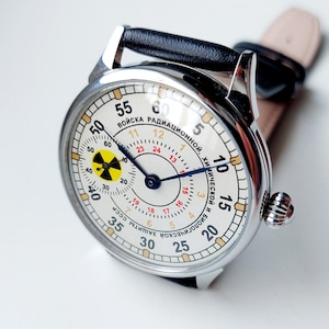 K&ouml;nnte beinhalten: Eine silberne Armbanduhr mit wei&szlig;em Zifferblatt und schwarzen Zeigern. Das Zifferblatt hat ein gelbes und schwarzes Strahlungssymbol und den Text "ВОЙСКА РАДИАЦИОННОЙ, ХИМИЧЕСКОЙ И БИОЛОГИЧЕСКОЙ ЗАЩИТЫ СССР" in kyrillischer Schrift.