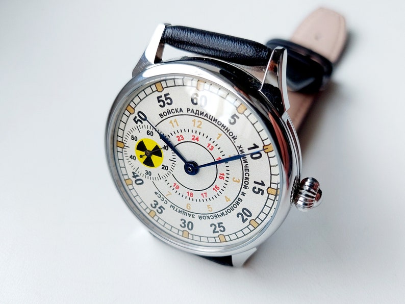 K&ouml;nnte beinhalten: Eine silberne Armbanduhr mit wei&szlig;em Zifferblatt und schwarzen Zeigern. Das Zifferblatt hat ein gelbes und schwarzes Strahlungssymbol und den Text "ВОЙСКА РАДИАЦИОННОЙ, ХИМИЧЕСКОЙ И БИОЛОГИЧЕСКОЙ ЗАЩИТЫ СССР" in kyrillischer Schrift.