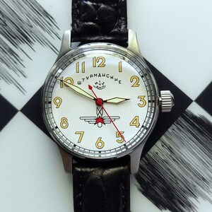 May include: A vintage wristwatch with a white face, gold numerals, and a black leather band. The watch features a red second hand and a silver case. The watch face has the text "Штурманские" and a logo.