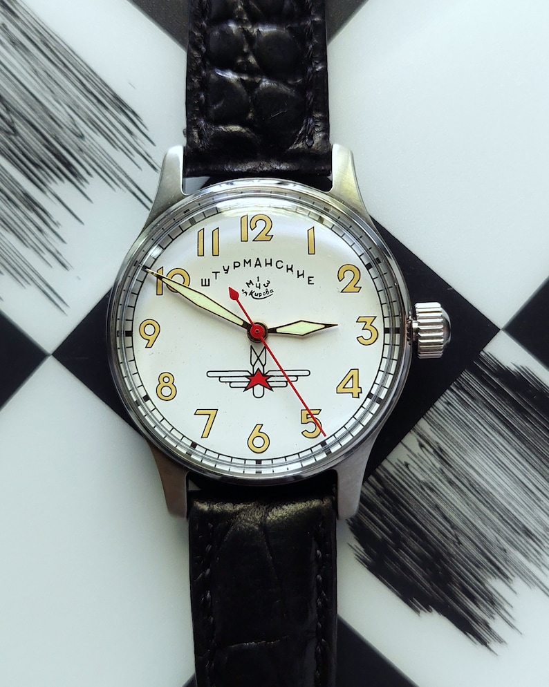 May include: A vintage wristwatch with a white face, gold numerals, and a black leather band. The watch features a red second hand and a silver case. The watch face has the text "Штурманские" and a logo.