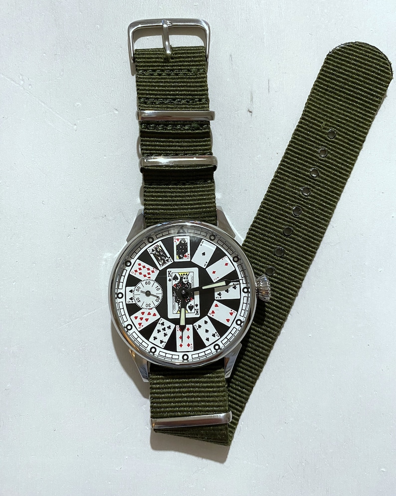 May include: A silver wristwatch with a green nylon strap. The watch face features a unique design with playing cards instead of numbers.