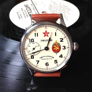 May include: A silver wristwatch with a white face and black hands. The watch face has red numbers and a red star with the text "КОМАНДИР" and "СМЕРТЬ ШПИОНАМ ЗАКАЗ ГРУ СССР".