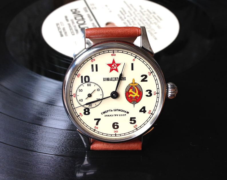 May include: A silver wristwatch with a white face and black hands. The watch face has red numbers and a red star with the text "КОМАНДИР" and "СМЕРТЬ ШПИОНАМ ЗАКАЗ ГРУ СССР".