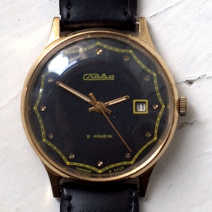 May include: A gold-tone wristwatch with a black face and a black leather band. The watch face has a gold-tone border with a yellow design and gold-tone hour markers. The watch face also has a date window at the 3 o'clock position. The watch face says "Слава" and "21 КАМЕНЬ" in Cyrillic.