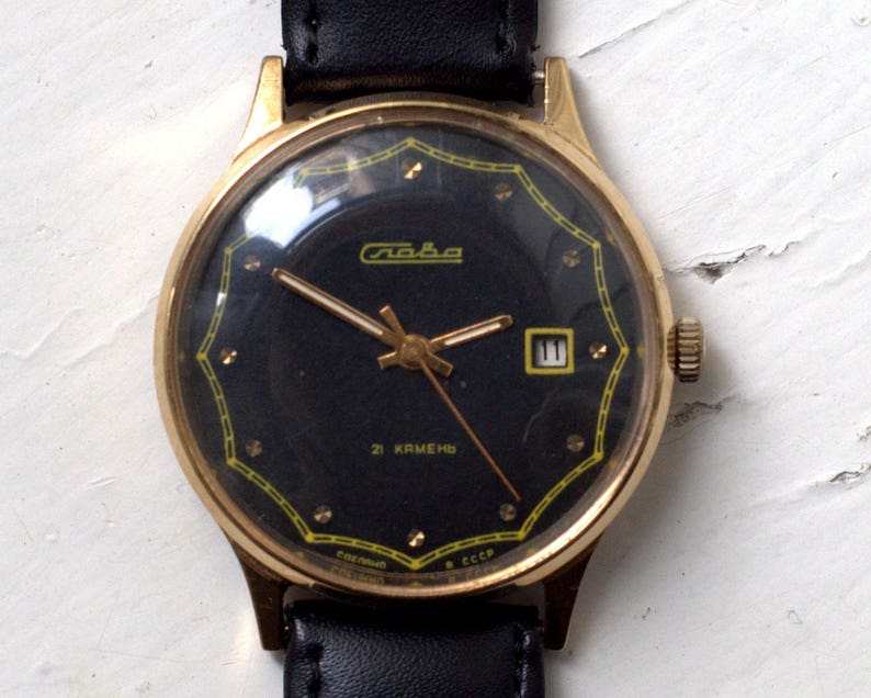 May include: A gold-tone wristwatch with a black face and a black leather band. The watch face has a gold-tone border with a yellow design and gold-tone hour markers. The watch face also has a date window at the 3 o'clock position. The watch face says "Слава" and "21 КАМЕНЬ" in Cyrillic.