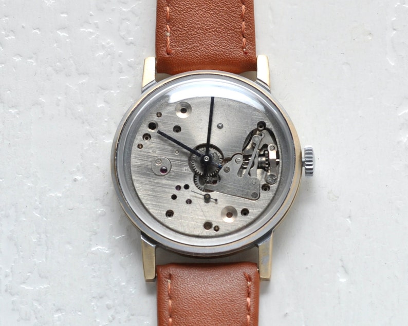 May include: A gold-tone wristwatch with a brown leather band. The watch face is clear and shows the inner workings of the watch.