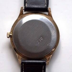 May include: A gold-tone wristwatch with a black leather band. The back of the watch is made of stainless steel and has the number "230" engraved on it.