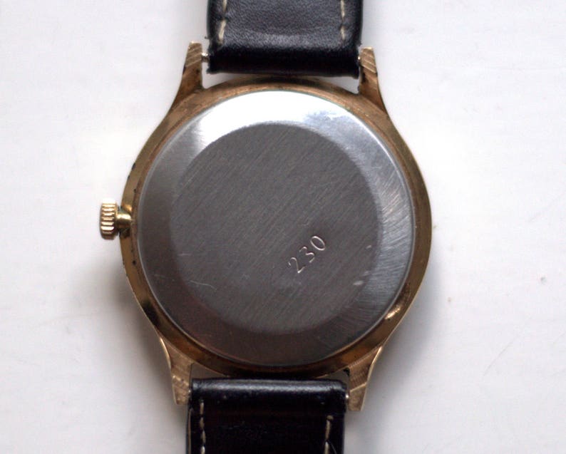 May include: A gold-tone wristwatch with a black leather band. The back of the watch is made of stainless steel and has the number "230" engraved on it.