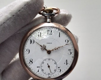 May include: A vintage Omega pocket watch with a white face, black numerals, and a subdial. The watch has gold-tone hands and a gold-tone crown. The watch is held by a gloved hand.