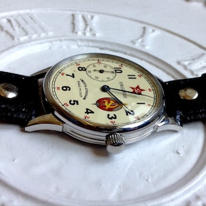 May include: A silver wristwatch with a white face and black leather band. The watch face has black Roman numeral hour markers, black hands, and a red star with a hammer and sickle emblem. The watch face also has the text "СССР" and "СДЕЛАНО В СССР" in Cyrillic script.