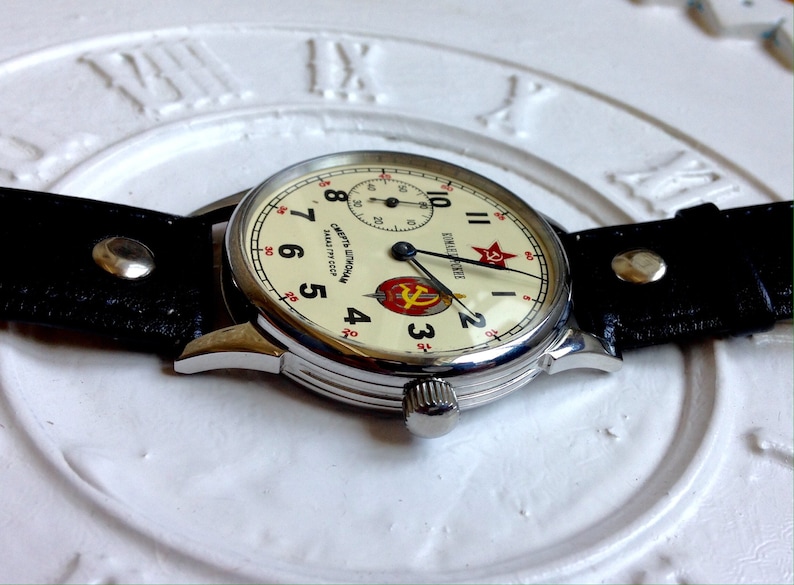 May include: A silver wristwatch with a white face and black leather band. The watch face has black Roman numeral hour markers, black hands, and a red star with a hammer and sickle emblem. The watch face also has the text "СССР" and "СДЕЛАНО В СССР" in Cyrillic script.