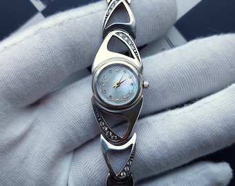 May include: A silver watch with a white face and a silver band. The band is made up of a series of linked triangles with small clear stones set into the edges.